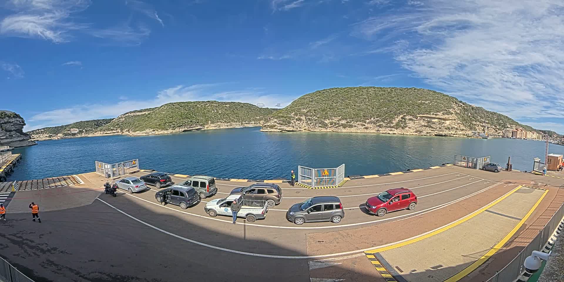 A ferry dock with several cars waiting to board, under a mostly sunny sky with a few clouds, overlooks a calm body of water surrounded by green hills and cliffs.