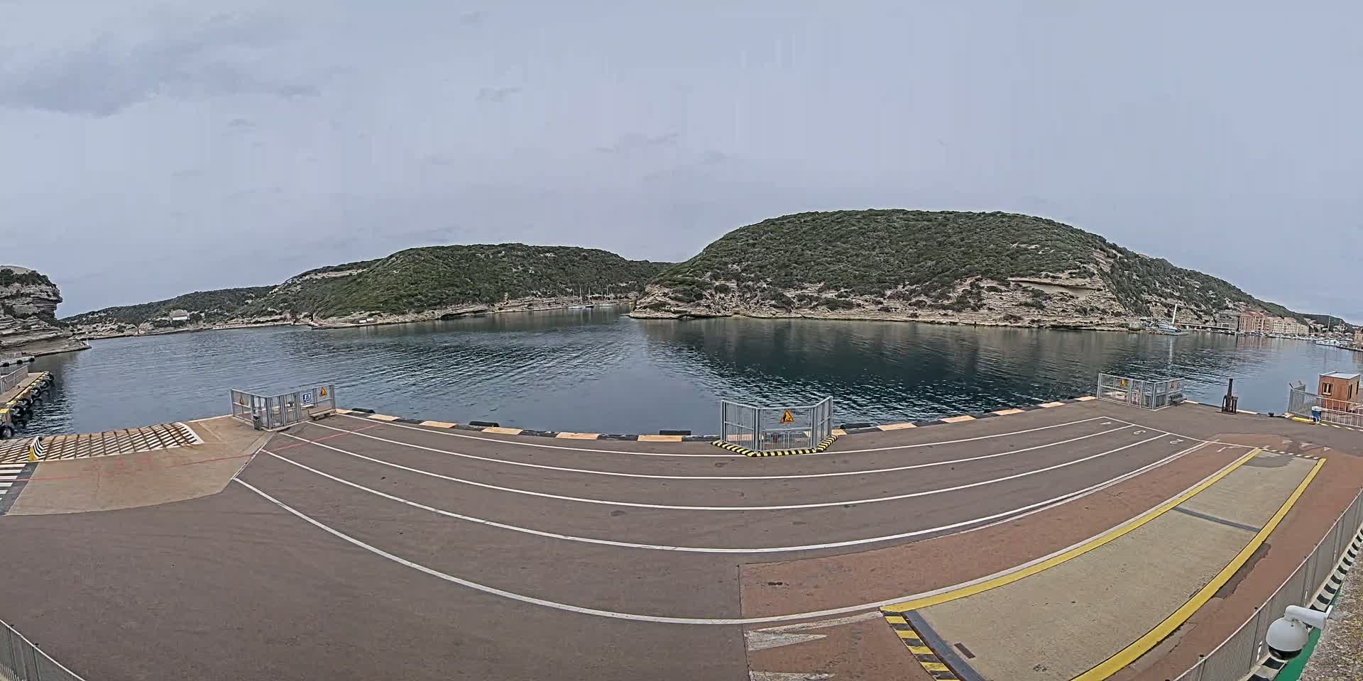 A ferry terminal with marked lanes overlooks a calm bay surrounded by green hills under an overcast sky.