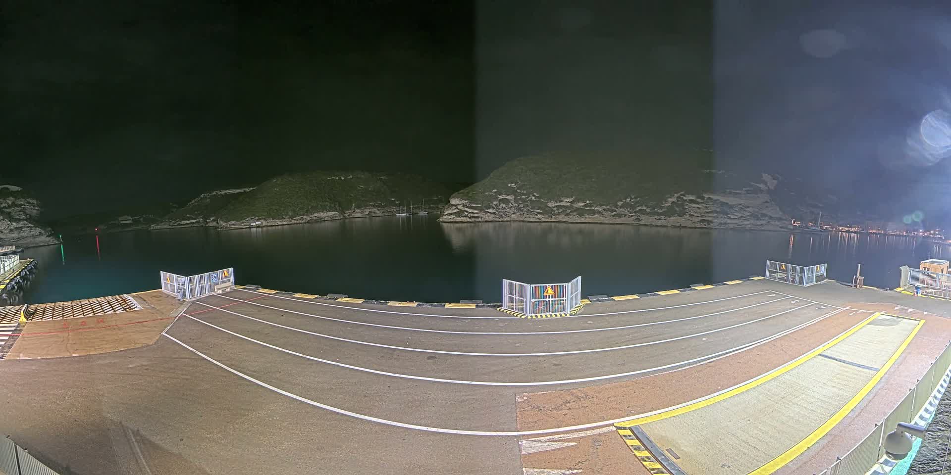 A nighttime view of an empty ferry dock at a calm bay, surrounded by dark hills under a cloudy sky.