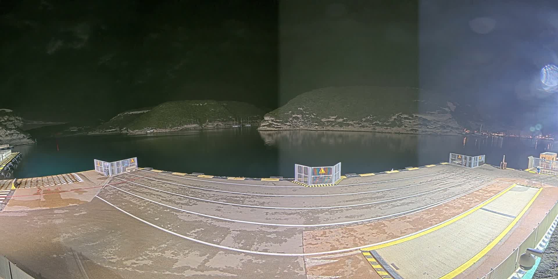 A nighttime view of an empty ferry dock with a calm body of water reflecting the dark hills and distant lights.