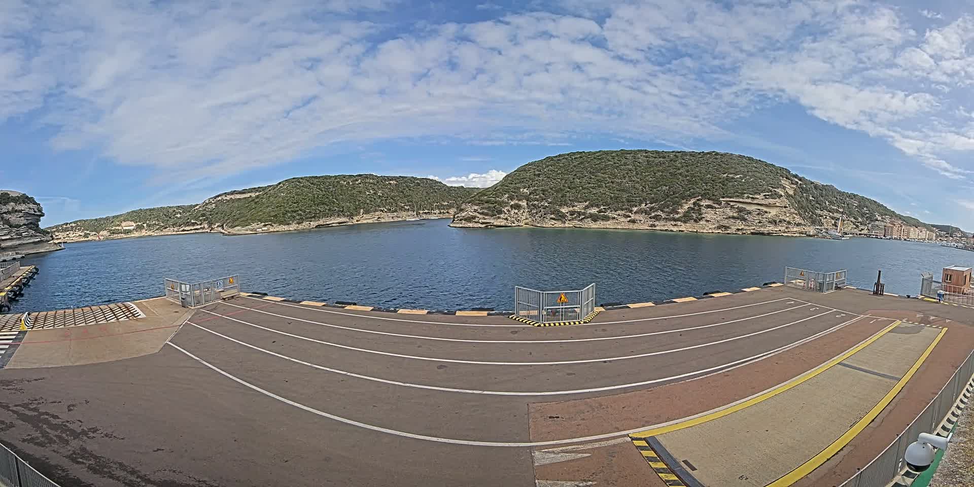 A ferry dock with marked lanes overlooks a calm body of water surrounded by green, rocky hills under a partly cloudy sky.