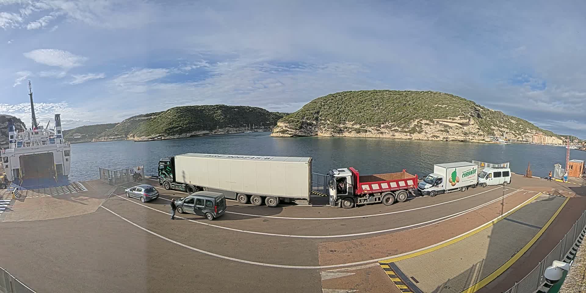 Several trucks, a van, and two cars are parked on a ferry terminal overlooking a calm body of water surrounded by hills under a partly cloudy sky.