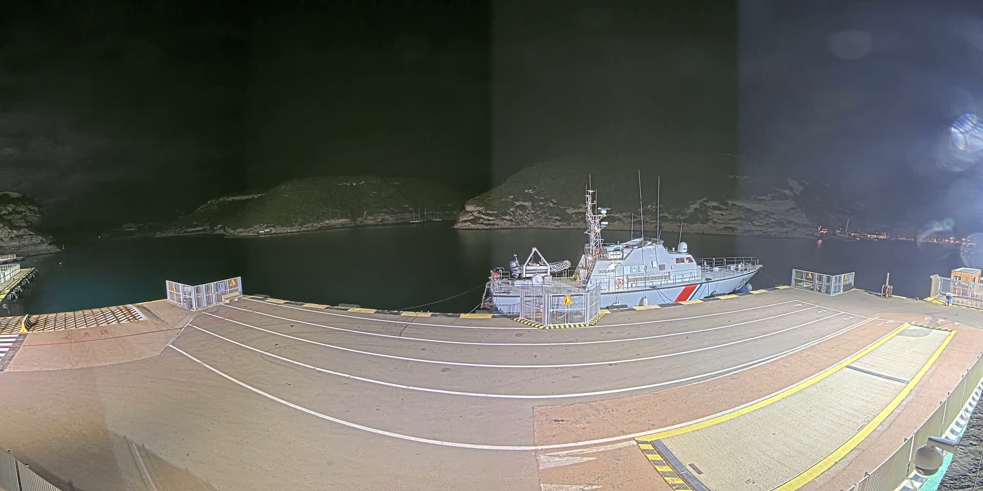A white patrol boat is docked at a nighttime harbor surrounded by dark hills under a mostly clear, dark sky.