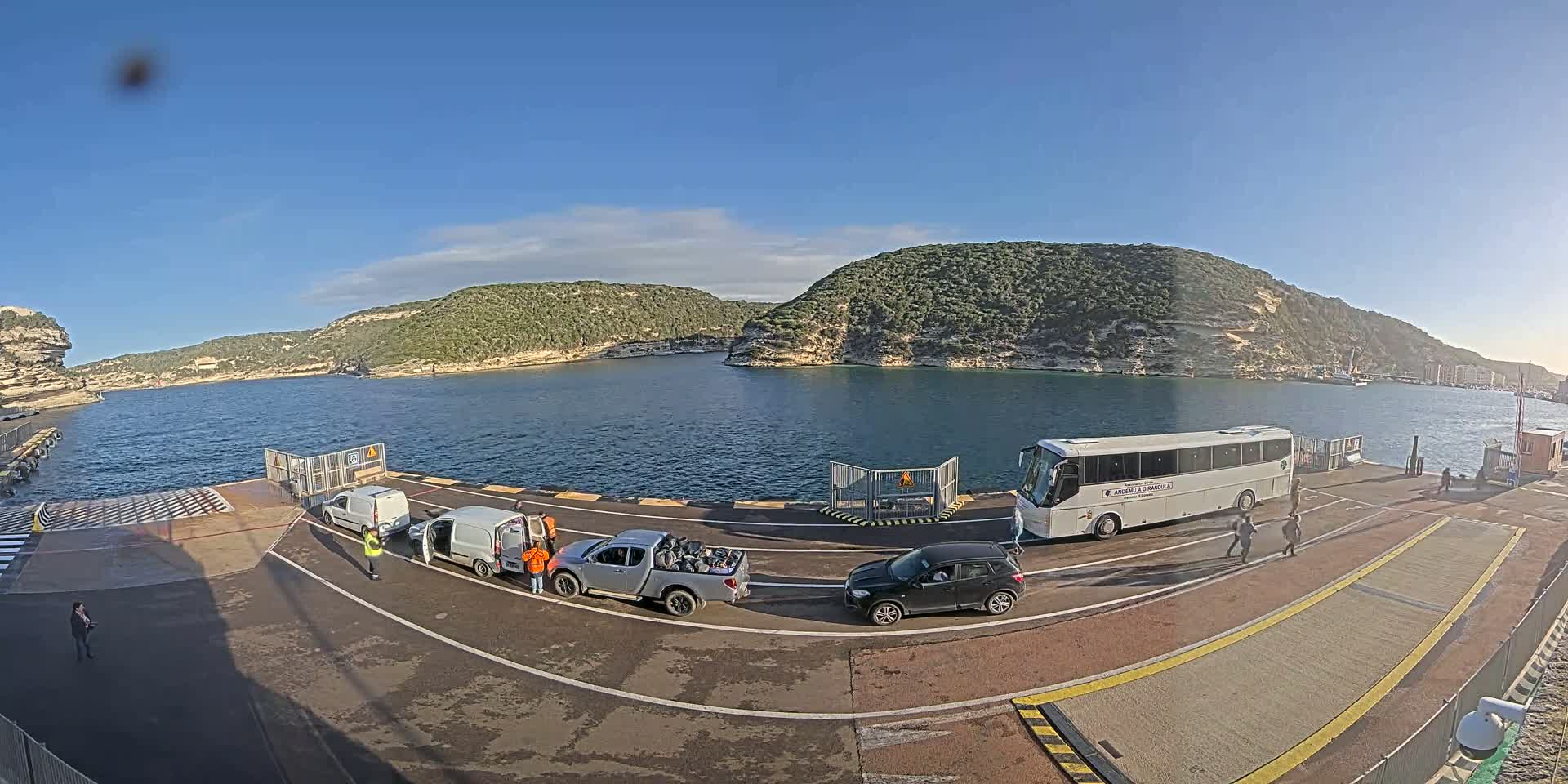 A ferry terminal on a sunny day with several vehicles, including a bus, van, pickup truck, and car, parked on the tarmac, overlooking a calm body of water surrounded by green hills.