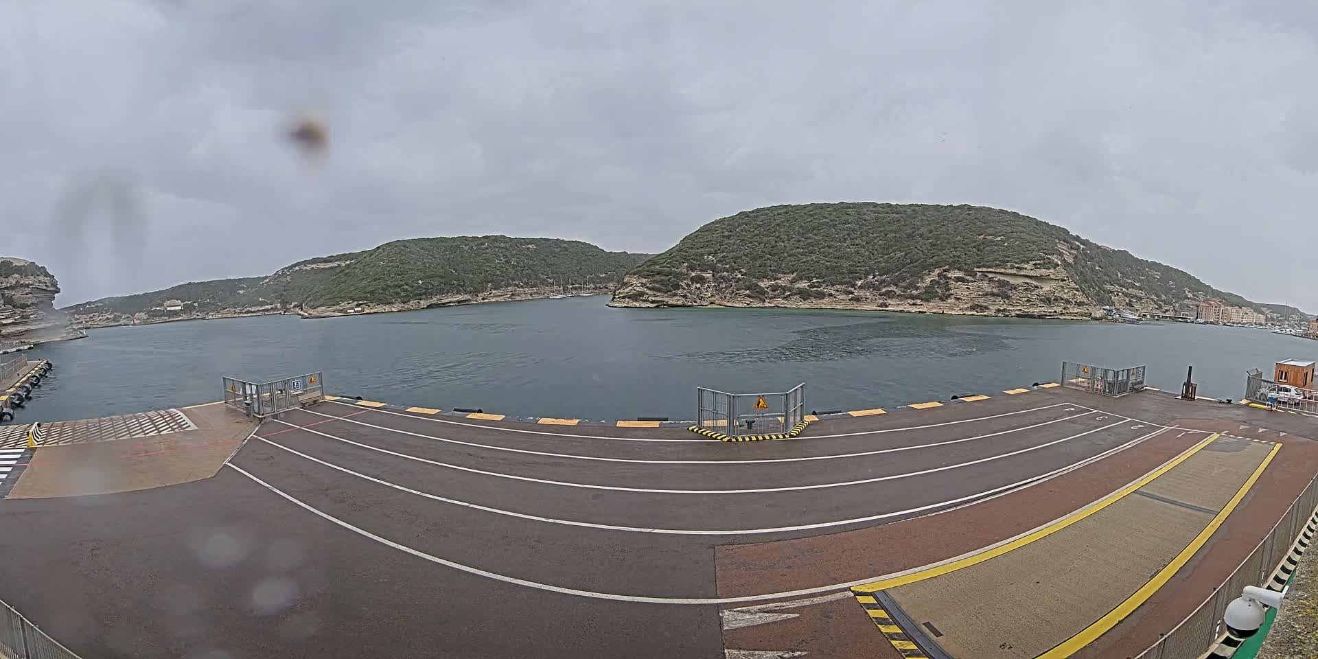 A ferry terminal with three lanes leading to a gate overlooks a calm body of water surrounded by green hills under an overcast sky.