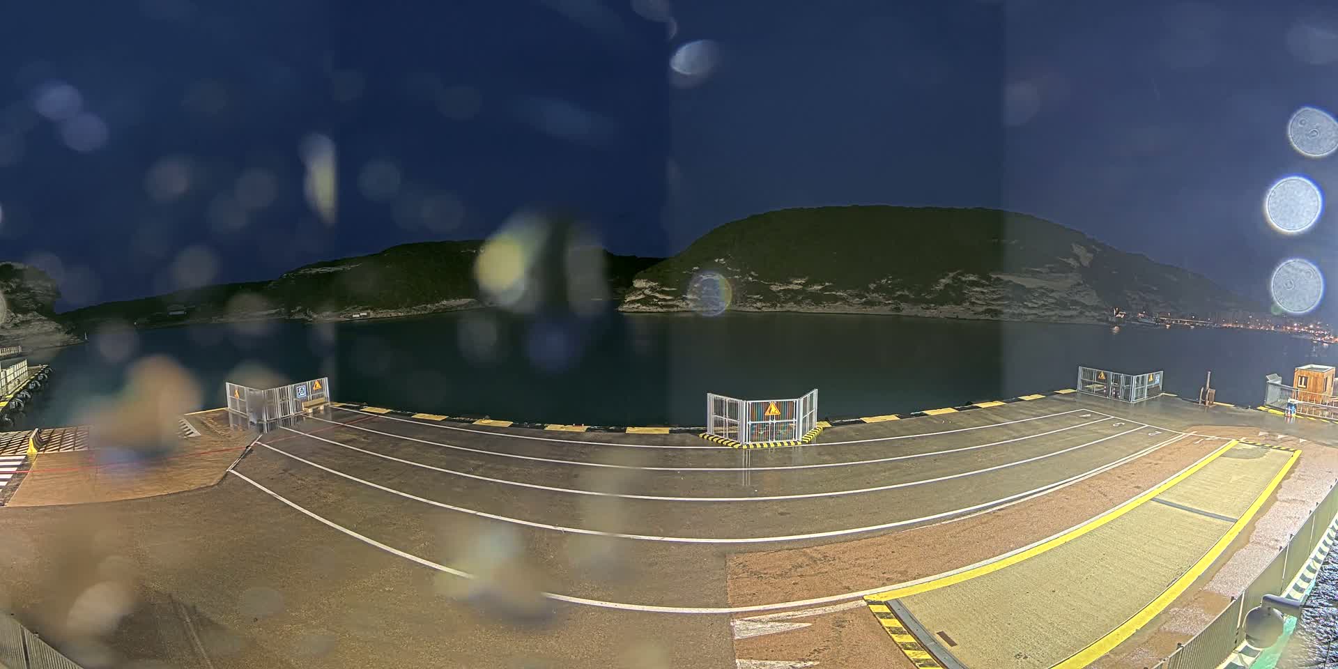 A rain-slicked ferry dock at night, with calm water and a dark, hilly shoreline visible in the background.