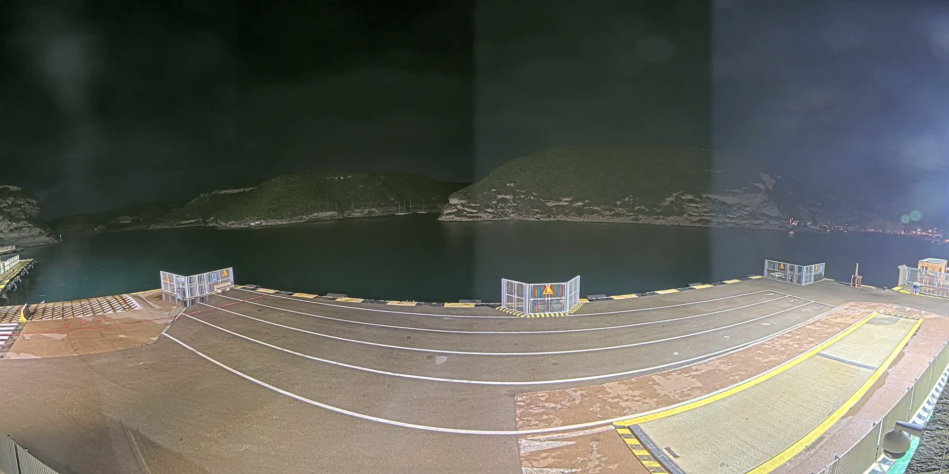 An empty ferry dock at night, situated in a calm bay surrounded by dark hills under a cloudy sky.
