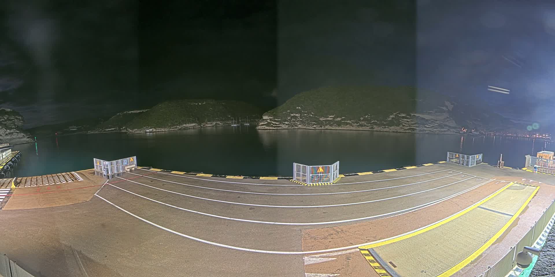 An empty ferry dock at night is illuminated, overlooking a dark, calm body of water surrounded by hills.