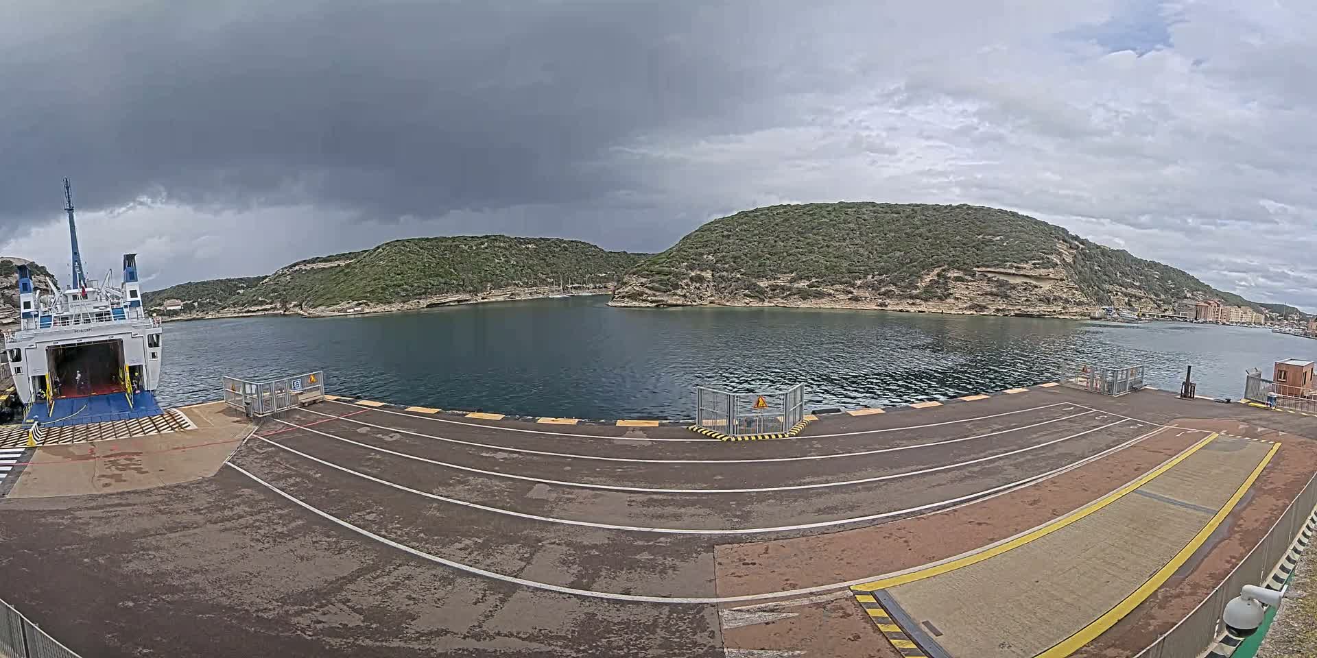 A ferry is docked at a port under a cloudy sky, with calm water and verdant hills in the background.