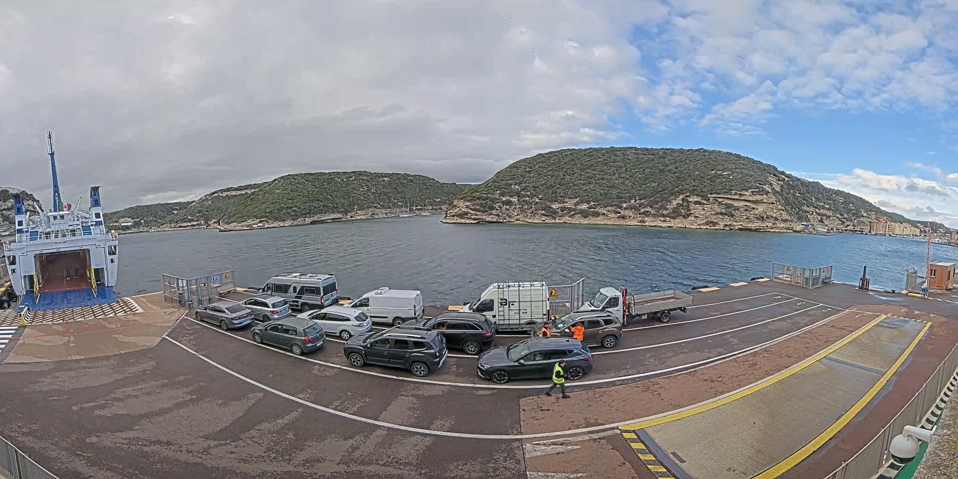 A ferry is docked at a port, with several cars and vans waiting to board under a partly cloudy sky.