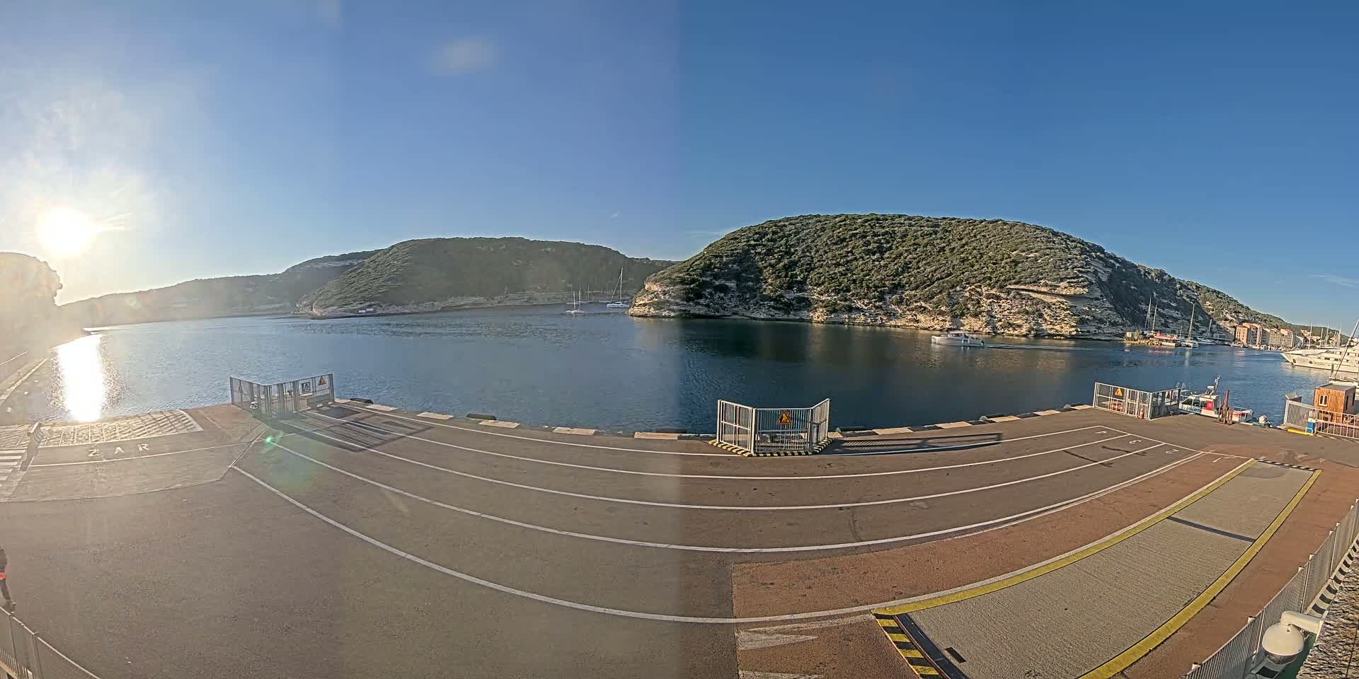 A sunny day reveals a harbor scene with calm water, several boats, and hills covered in green vegetation.