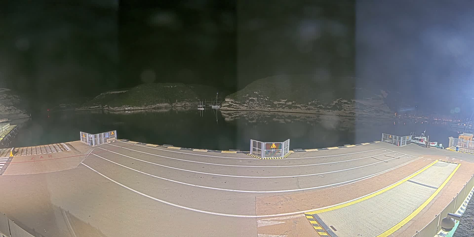 A nighttime view of a ferry dock with several boats in a calm, dark body of water surrounded by hills.