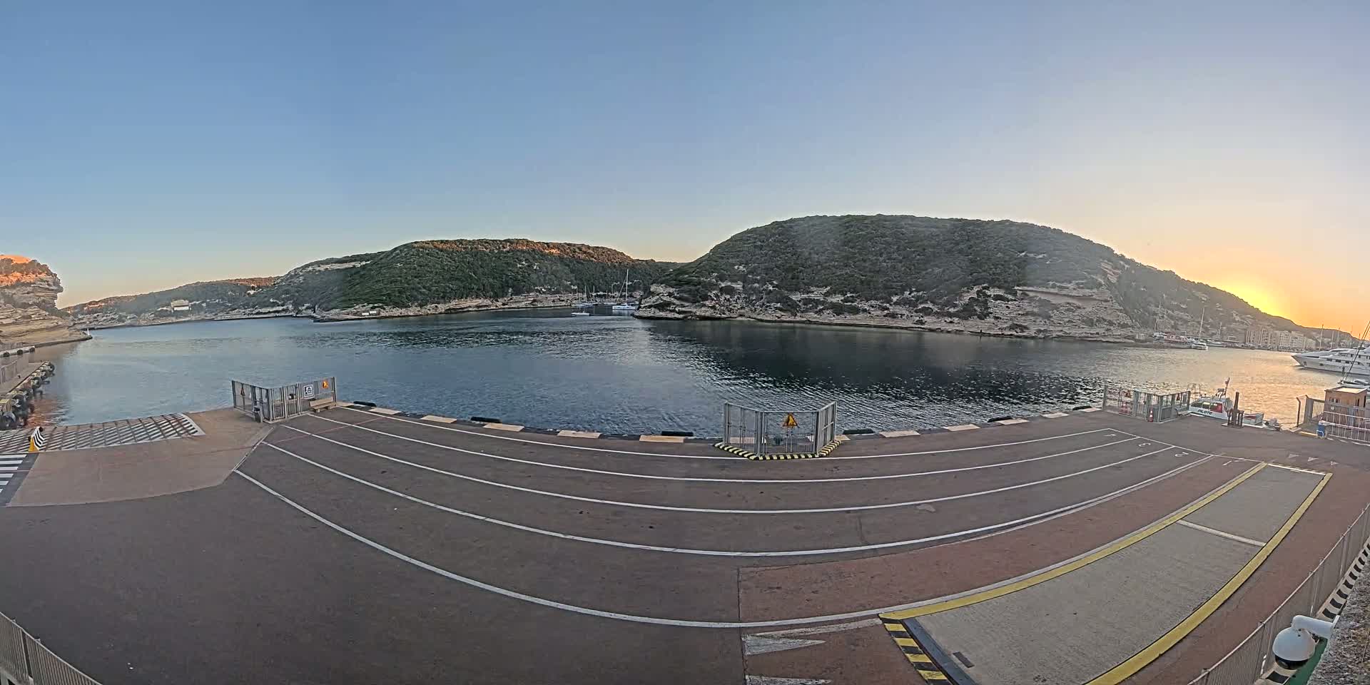 A calm harbor at sunset is viewed from a ferry dock, with several boats visible in the water and hills covered in green vegetation surrounding the water.