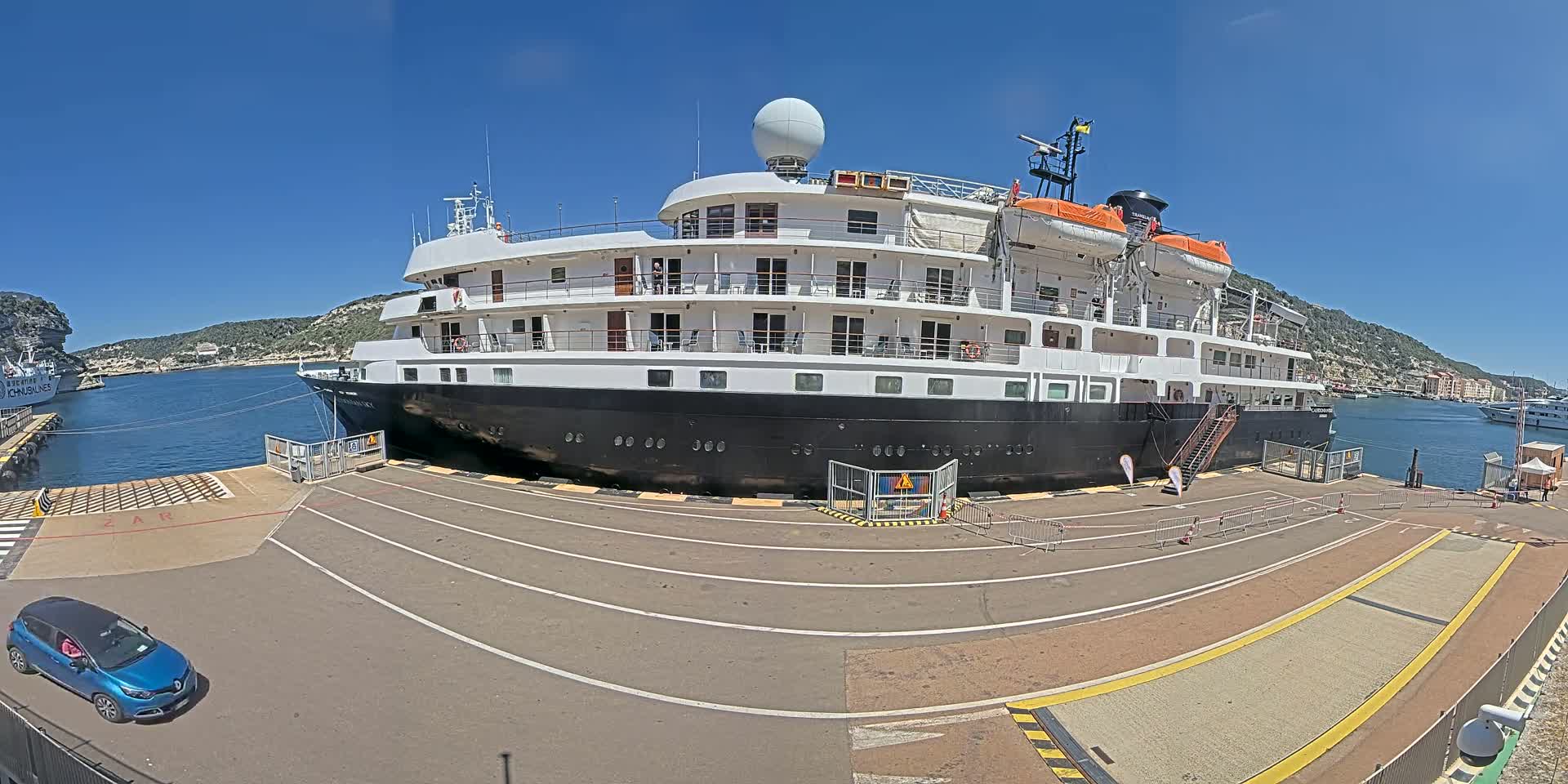 A large cruise ship is docked at a port on a sunny day, with a small blue car parked nearby.