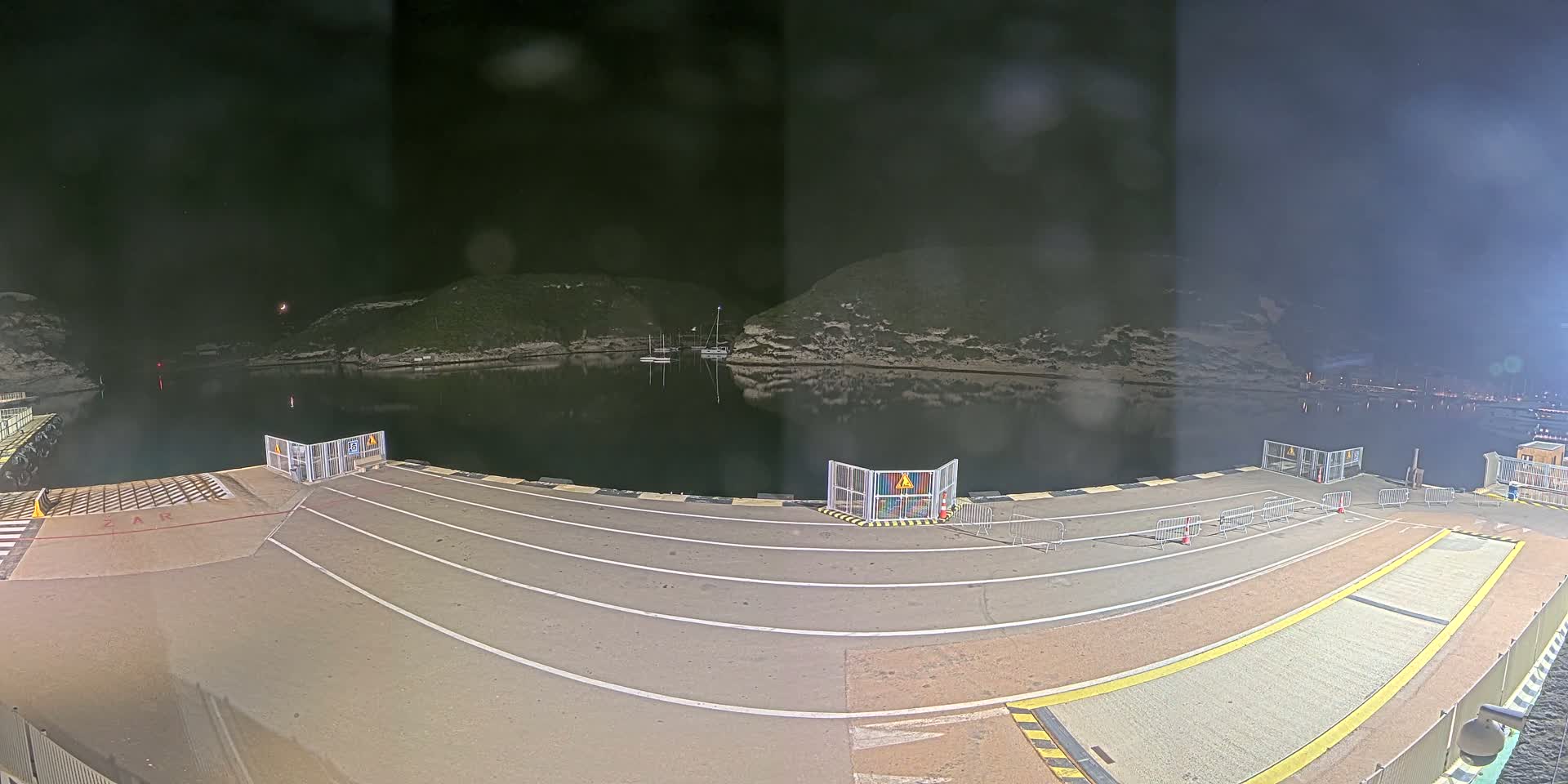 A nighttime view of an empty ferry terminal at a calm, dark-water harbor nestled between hills.