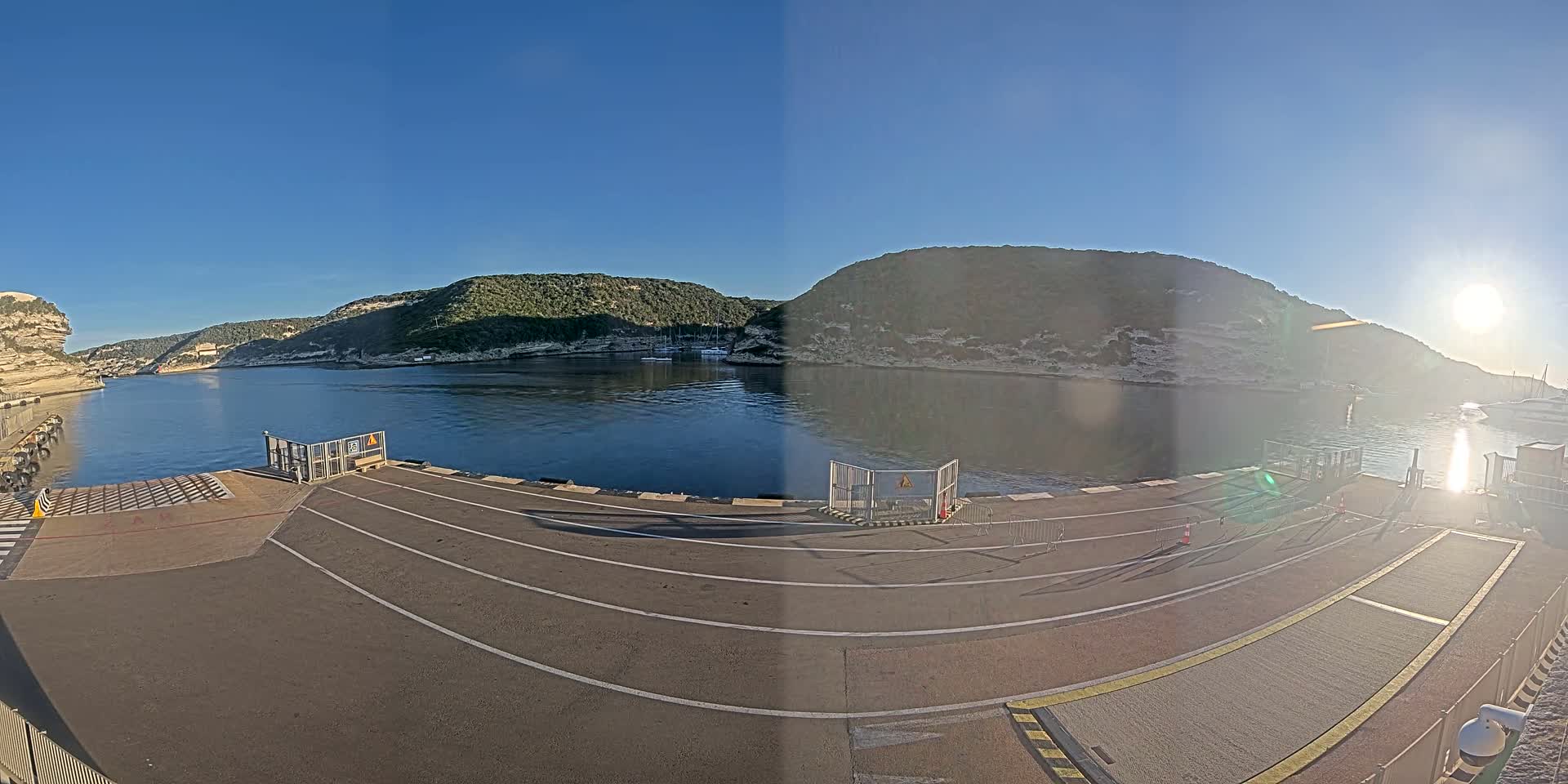 A ferry dock with marked lanes is situated on a calm body of water, surrounded by hills under a clear, sunny sky.
