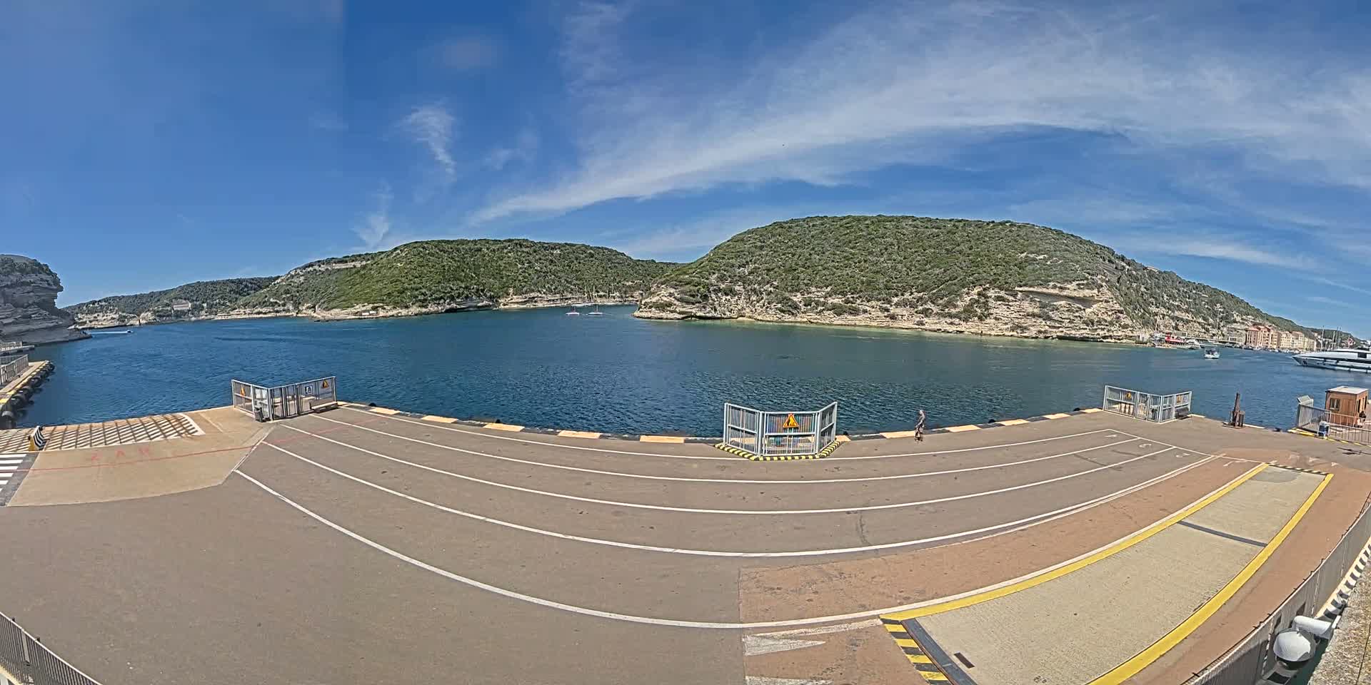 A ferry dock with marked lanes overlooks a calm bay surrounded by green hills under a mostly sunny sky.
