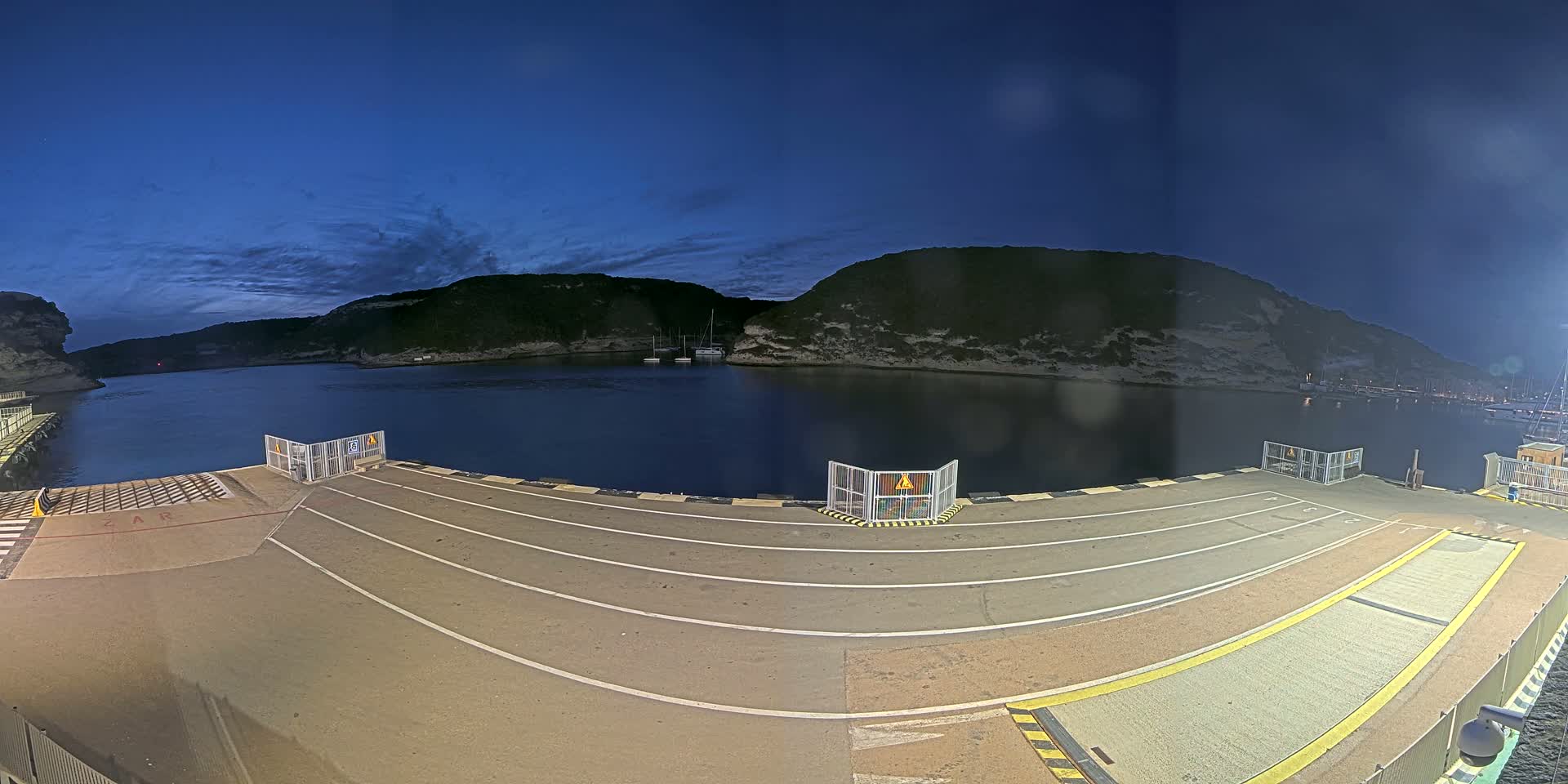 An empty ferry dock at night under a clear, dark blue sky, with calm water and hills visible in the background.