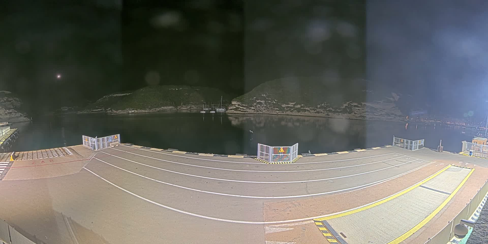 A nighttime view of an empty ferry dock overlooking a calm, dark body of water surrounded by low-lying hills.