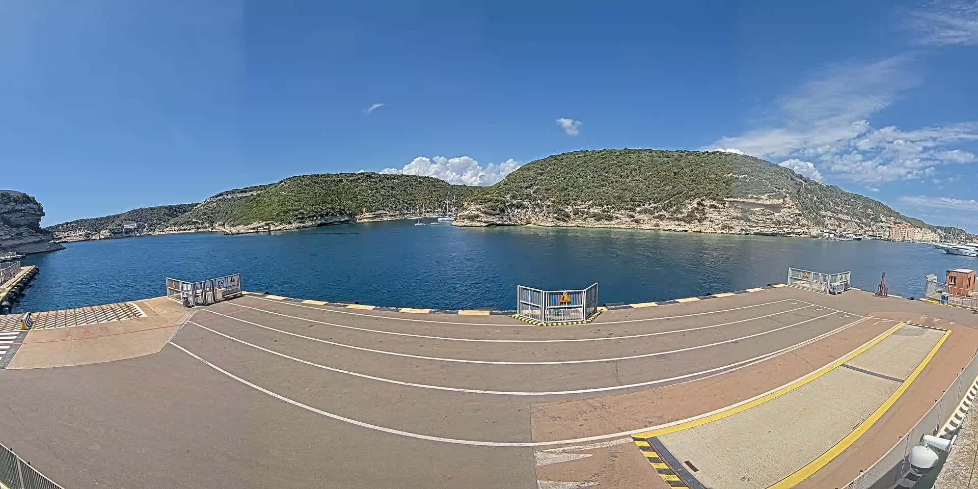A ferry dock with marked lanes overlooks a calm bay surrounded by green, rocky hills under a mostly sunny sky.