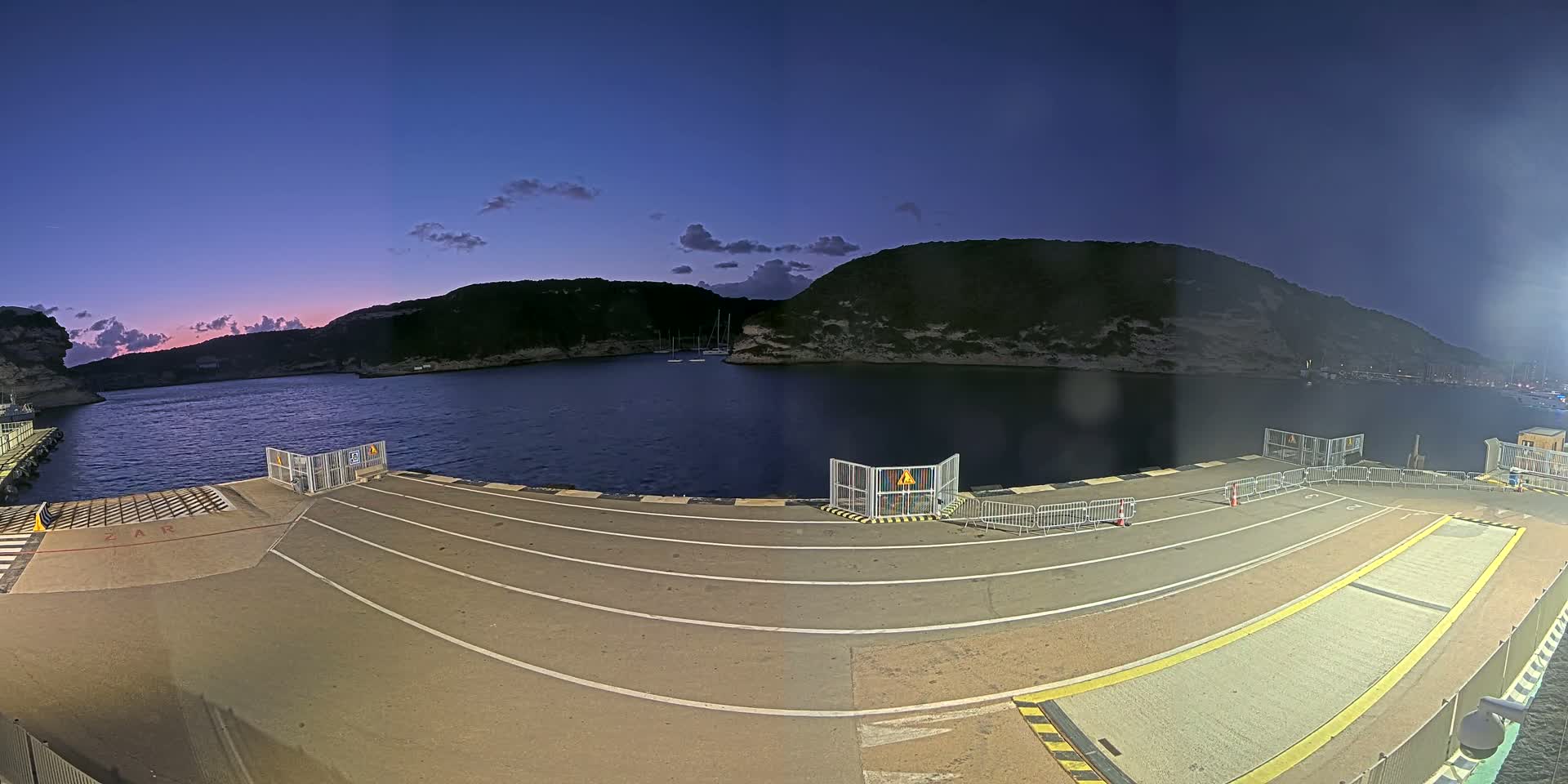 An empty ferry terminal at dusk overlooks a calm bay surrounded by dark, rocky hills under a twilight sky.