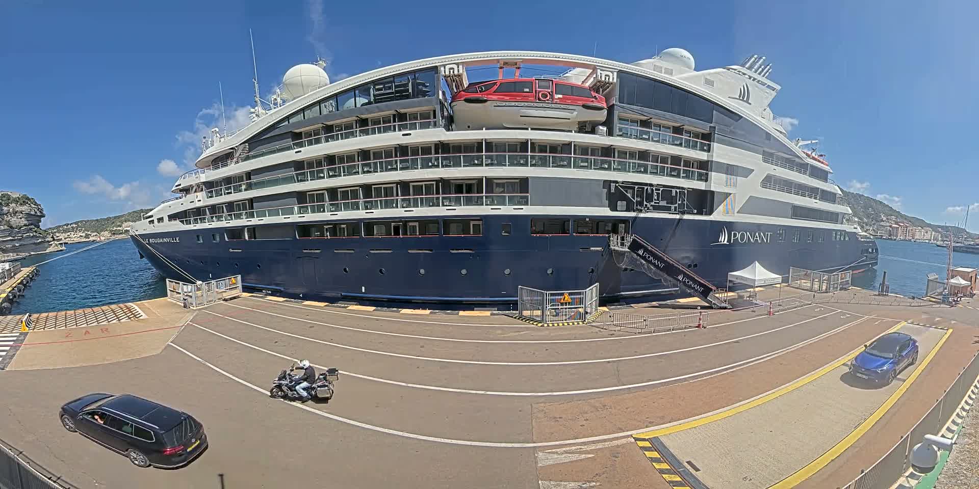 A large cruise ship is docked at a port on a sunny day, with two cars and a motorcycle visible on the dock.