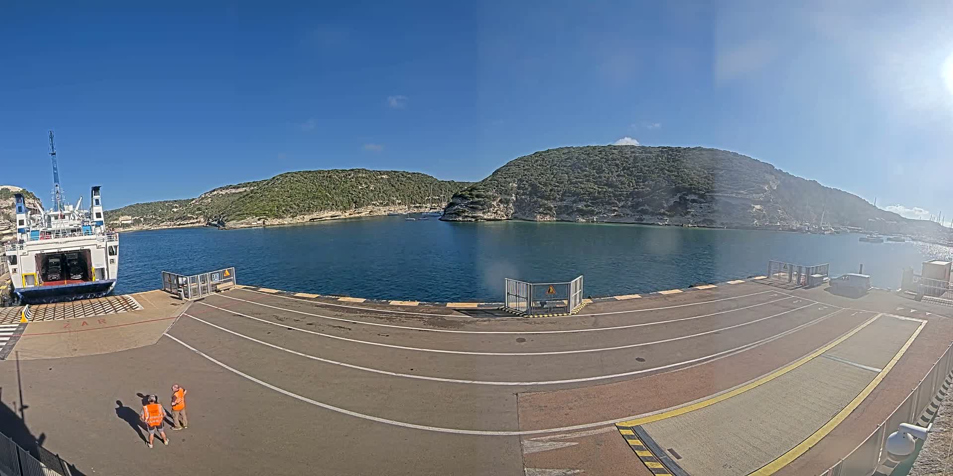 A ferry is docked at a port on a sunny day, with two people standing on the dock and green hills surrounding a calm body of water.