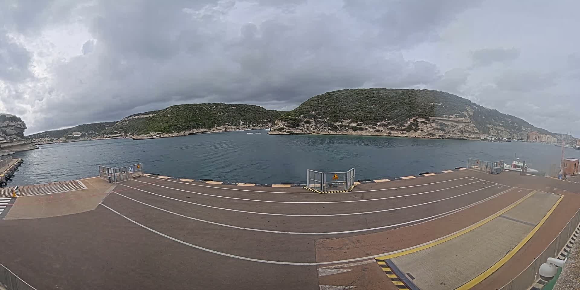 A ferry dock with a few boats in a calm bay surrounded by green, rocky hills under an overcast sky.
