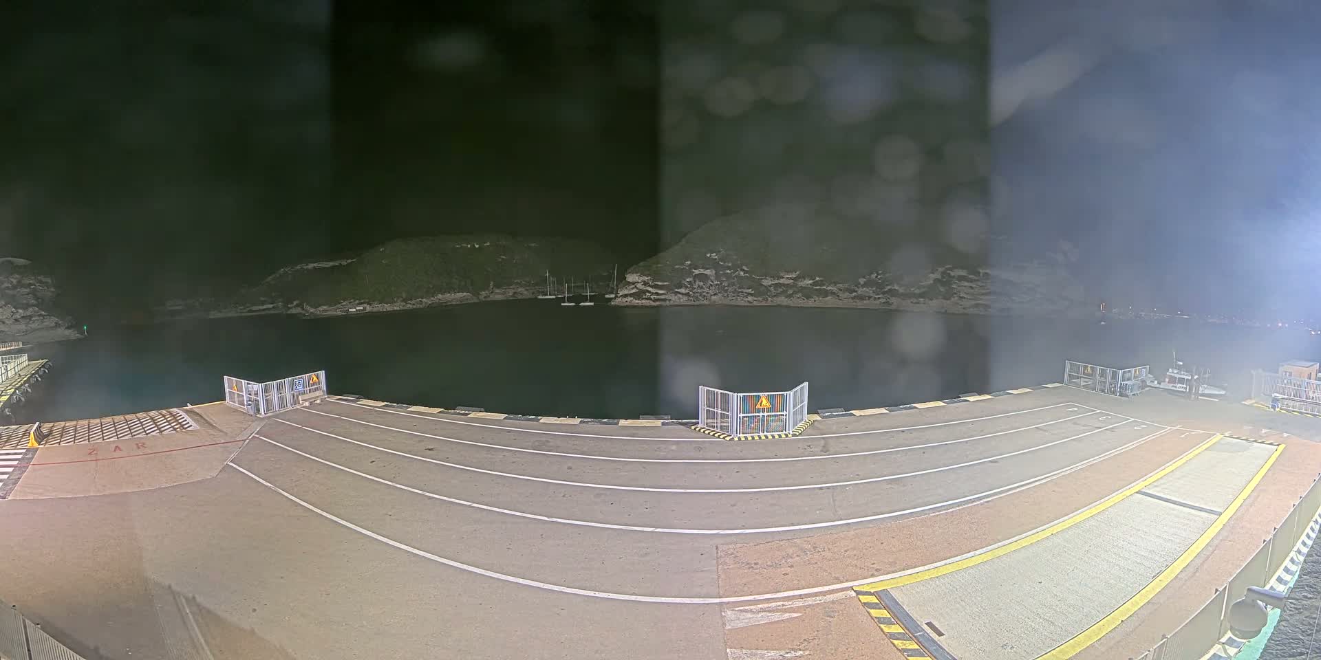 A nighttime view of a harbor's empty dock and calm water, with a few boats visible in the distance.