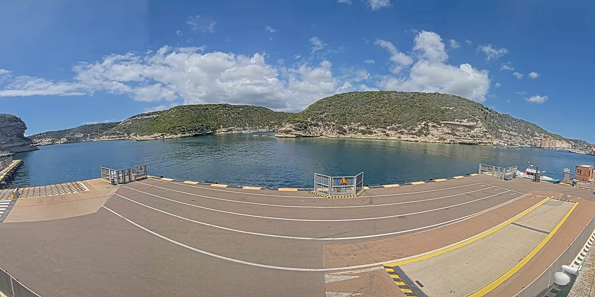 A ferry terminal's empty loading area with marked lanes overlooks a calm bay surrounded by green hills under a partly cloudy sky.