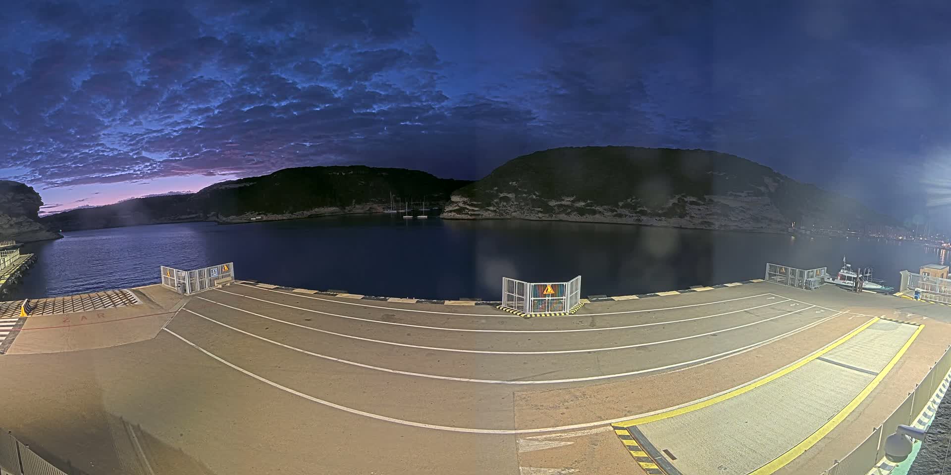 A ferry dock at twilight under a partly cloudy sky overlooks a calm body of water nestled between dark hills.