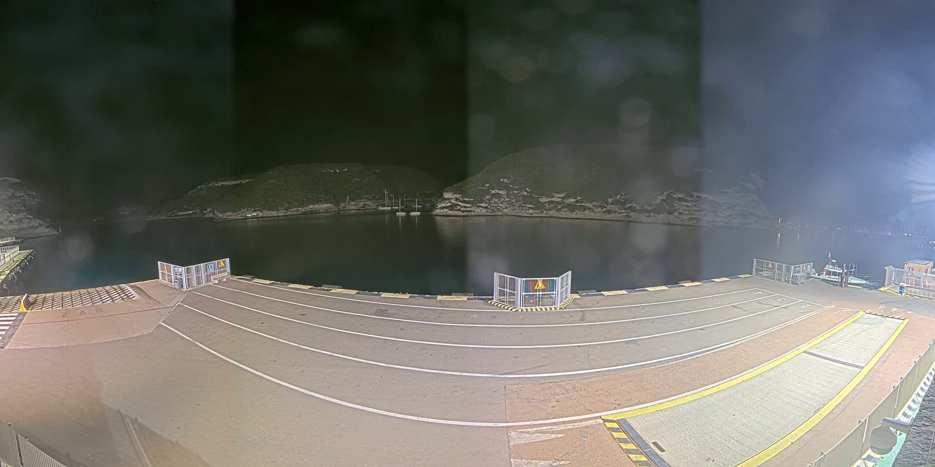 A nighttime view of a ferry dock with several boats moored in a calm, dark-colored body of water surrounded by hills.