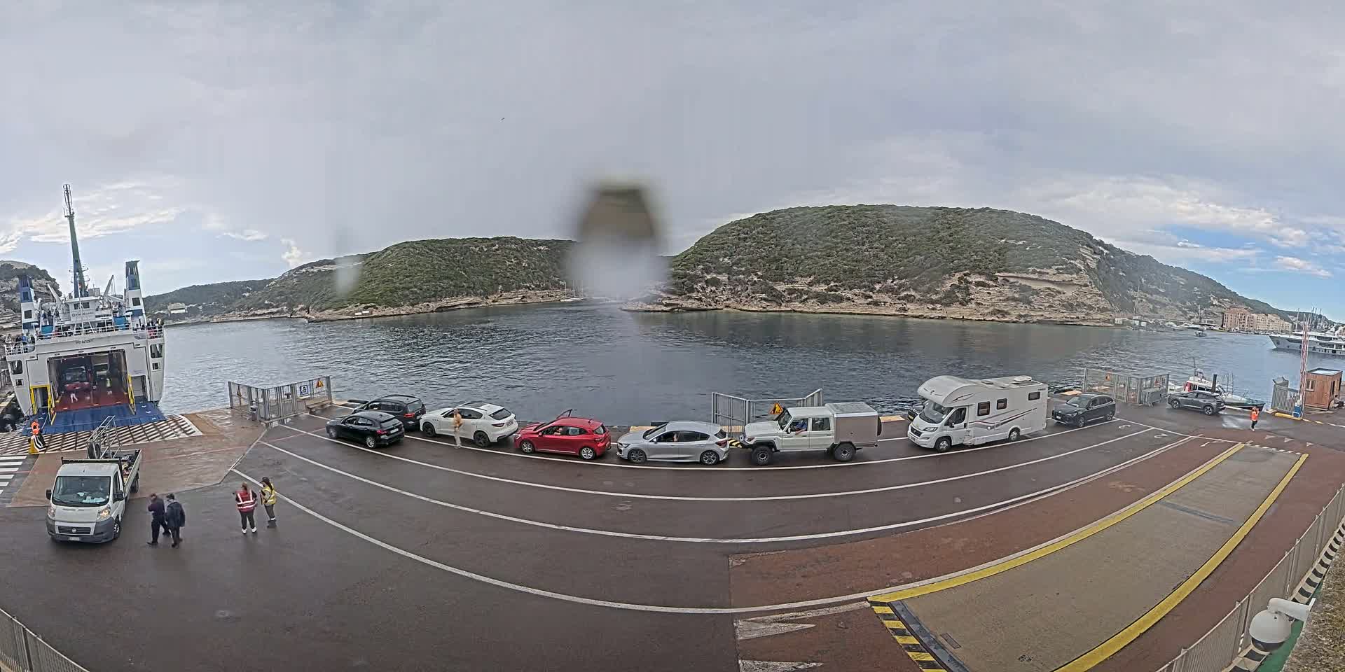 A ferry is docked at a port on a cloudy day, with several cars and a campervan lined up on the dock awaiting embarkation.