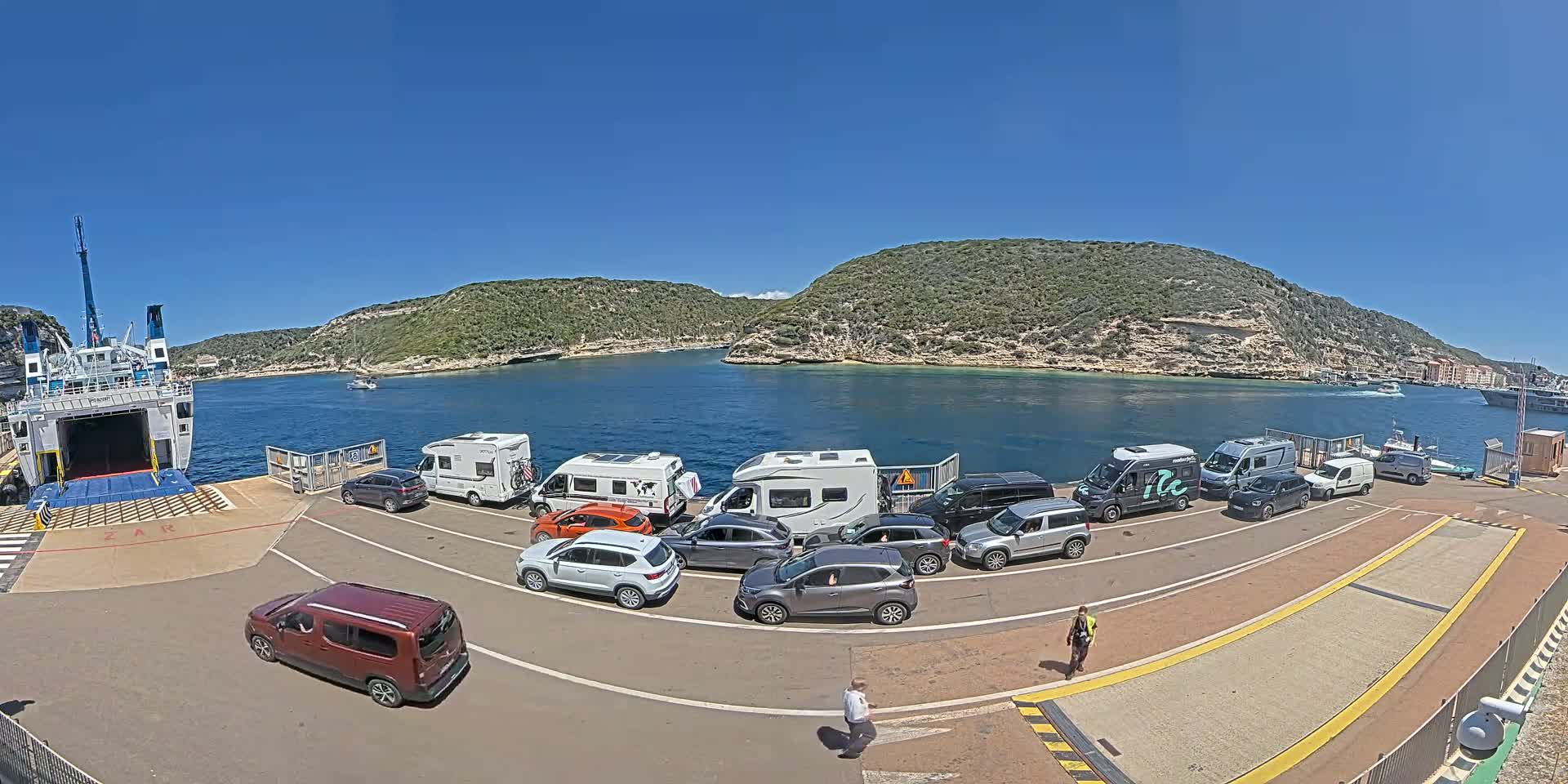 A ferry is docked at a port on a sunny day, with several cars and RVs lined up to board, situated in a calm bay surrounded by green hills.