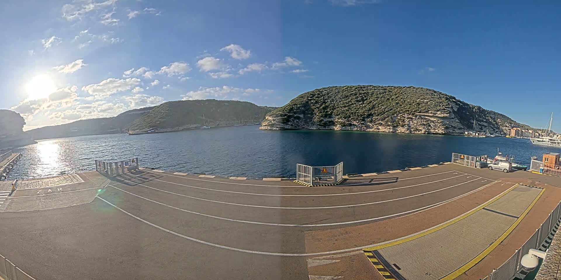 A sunny day at a harbor shows a paved area with vehicle lanes leading to a ferry dock, overlooking a calm bay surrounded by green hills.