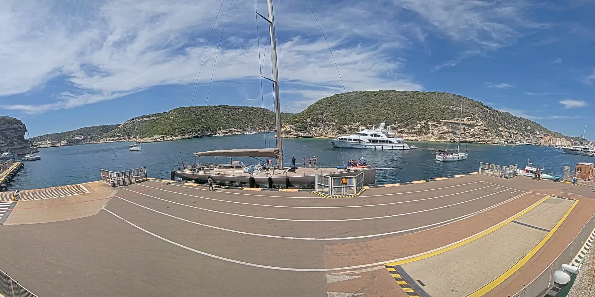 A large sailboat is docked at a marina on a sunny day with partly cloudy skies, surrounded by other boats and hills covered in green vegetation.
