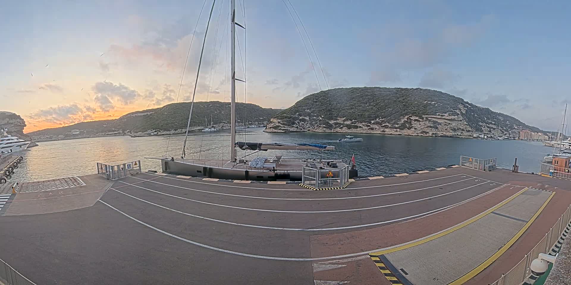 A large sailboat is docked at a marina during sunset, with calm water and hills in the background.