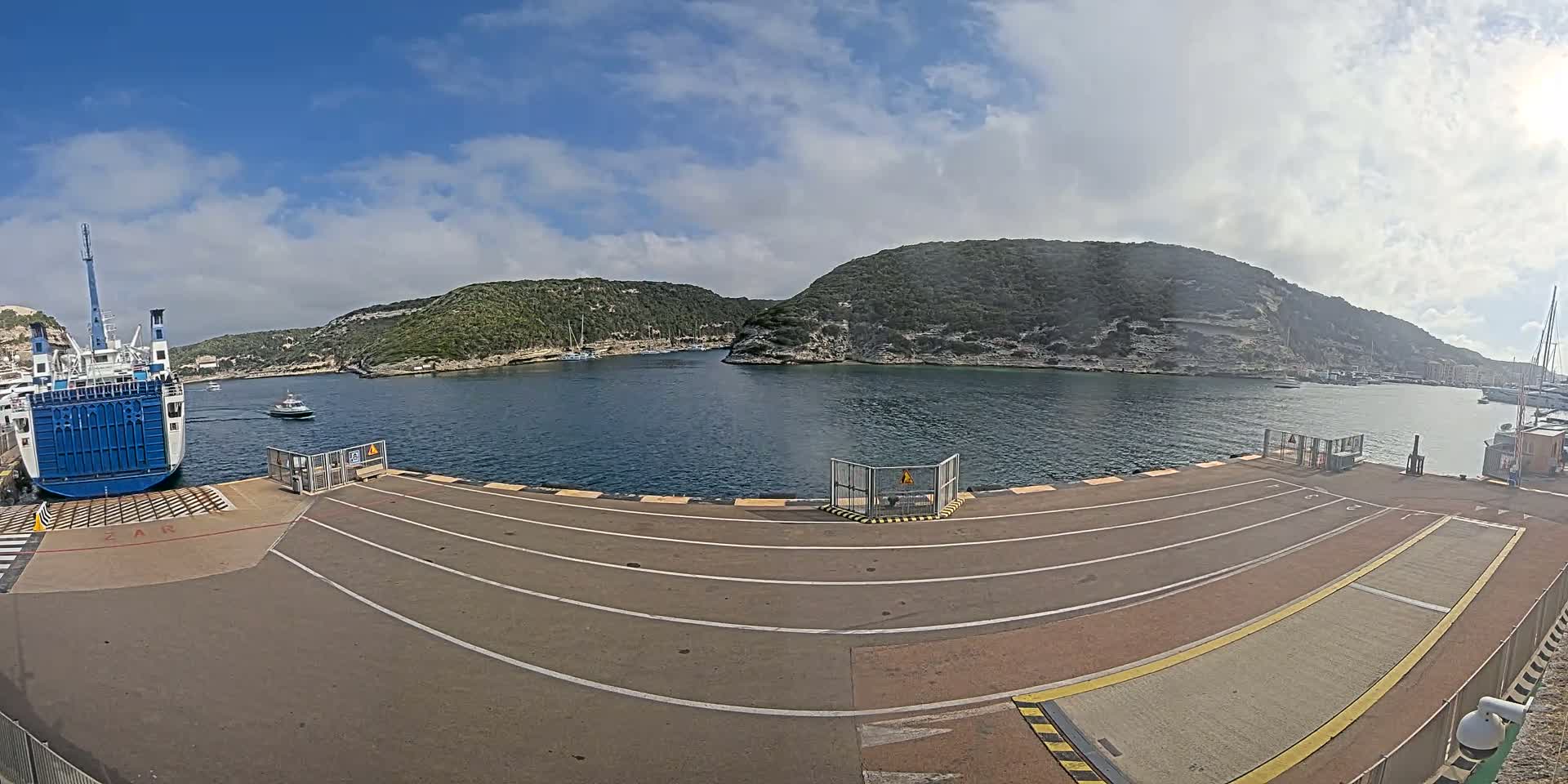 A ferry is docked at a port, with calm water and partly cloudy skies, surrounded by green hills.