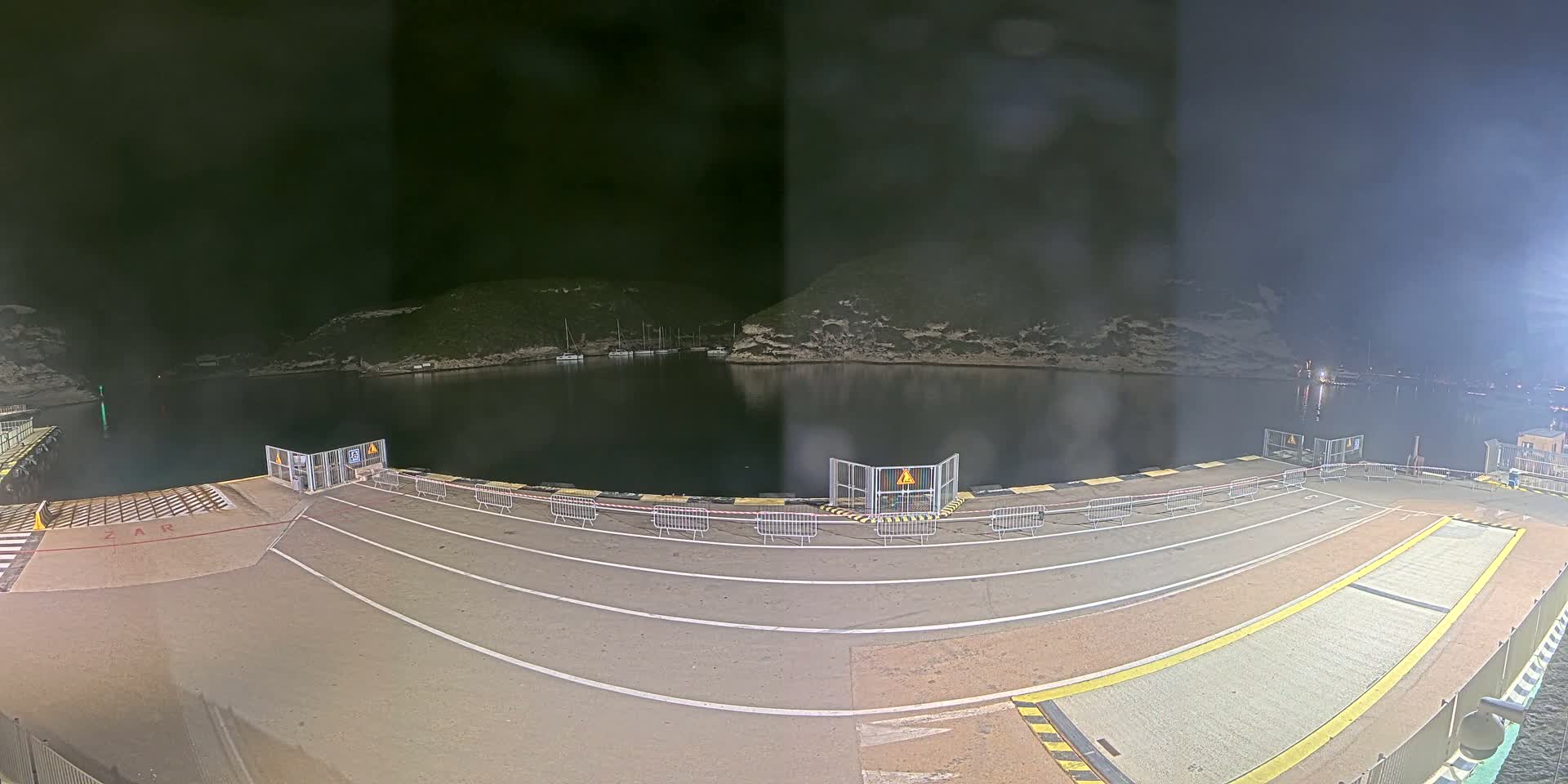 At night, an empty ferry dock is shown with a calm body of water and hills in the background.