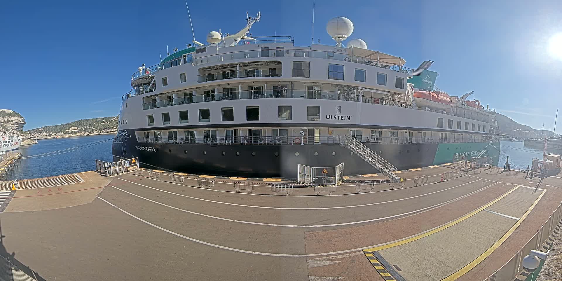A large cruise ship is docked at a port on a sunny day, with another smaller vessel visible in the background.