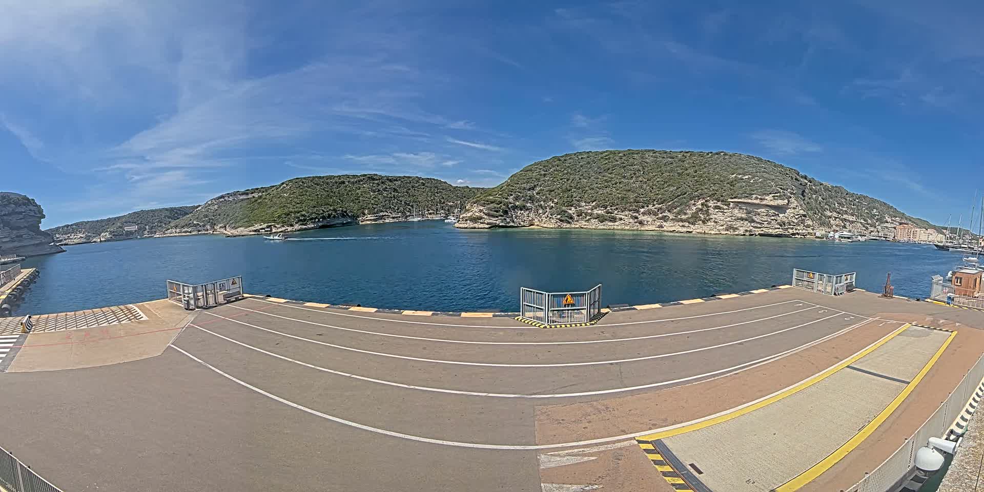 A ferry terminal's empty asphalt loading area overlooks a calm bay surrounded by rocky, green hills under a clear blue sky.