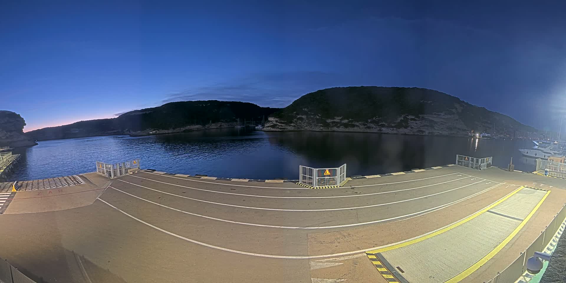 An empty ferry dock at twilight overlooks a calm bay bordered by dark, rocky hills under a clear, dark blue sky.