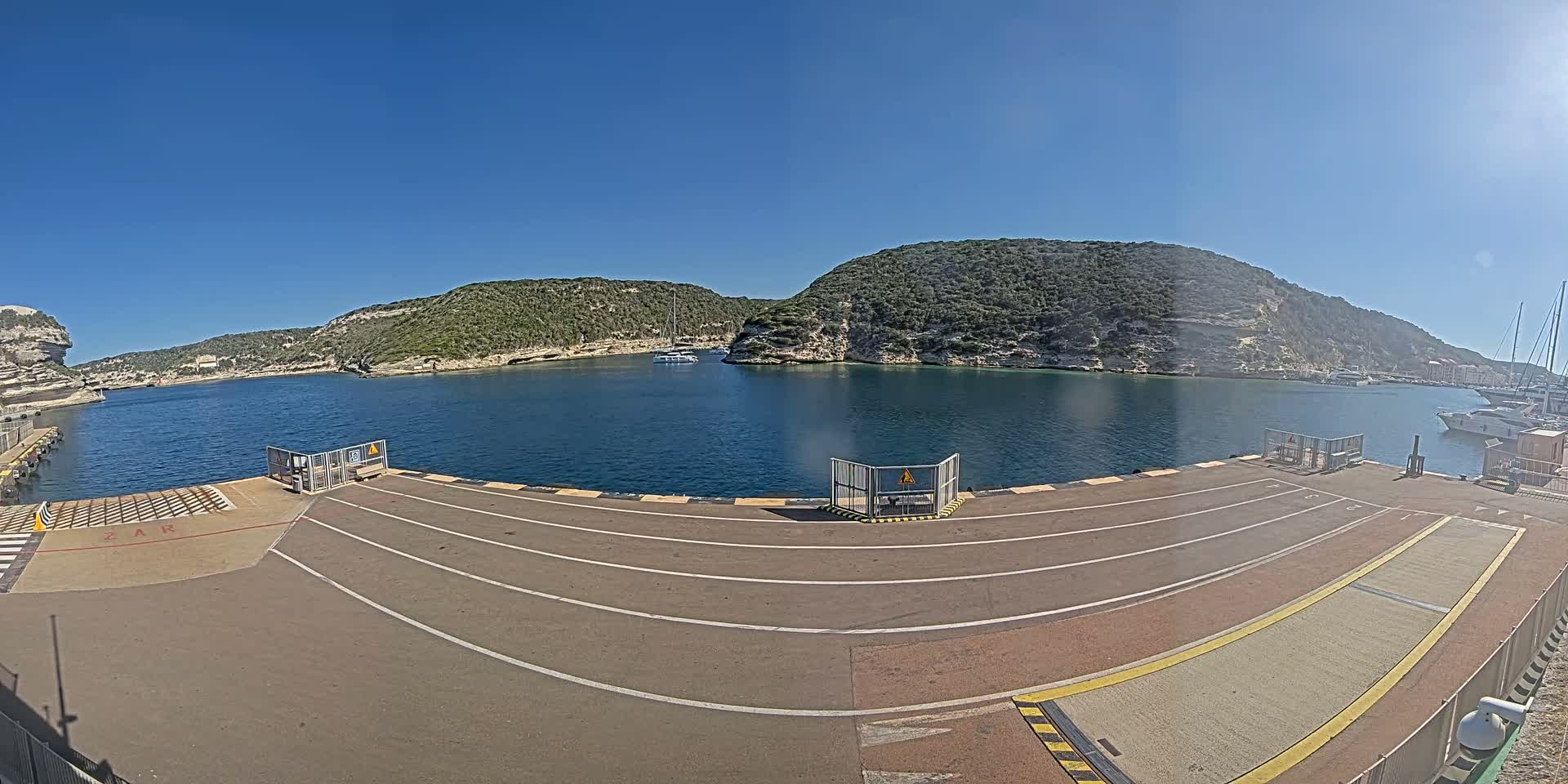 A ferry dock with marked lanes overlooks a calm bay surrounded by green hills under a clear blue sky.