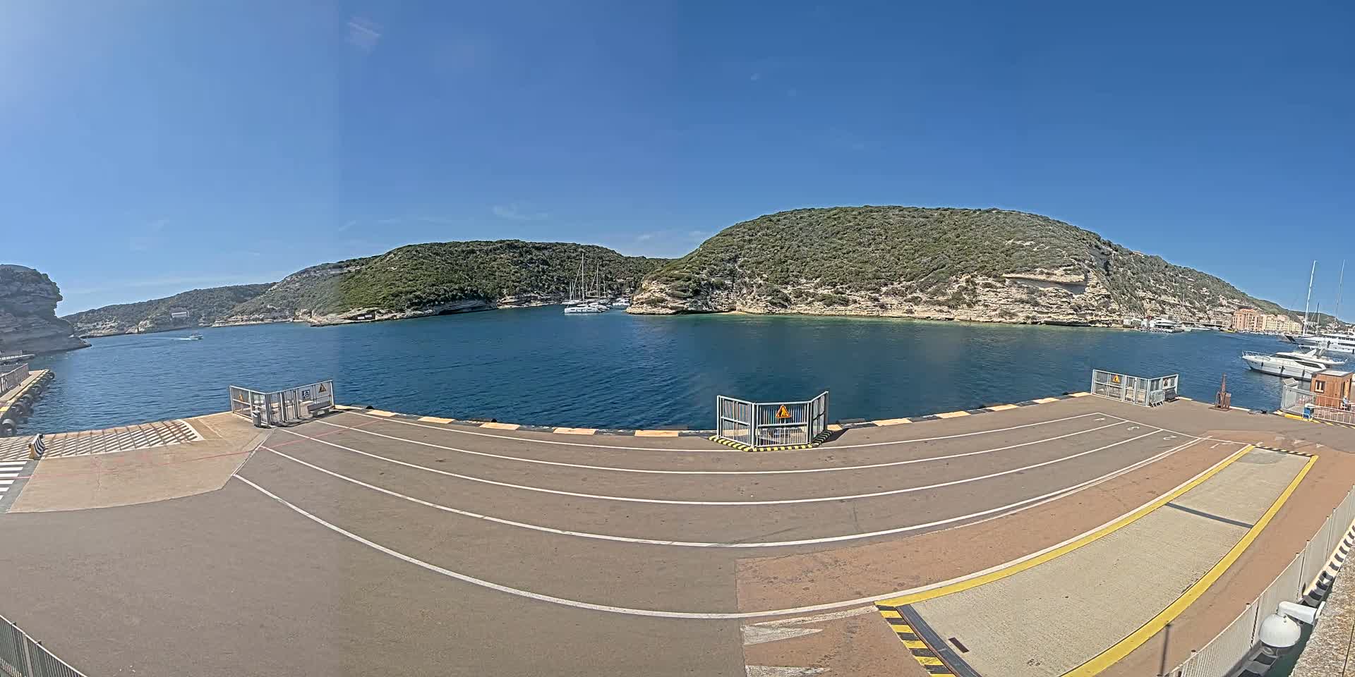 A ferry dock with marked lanes overlooks a calm bay surrounded by green, rocky hills under a clear blue sky.