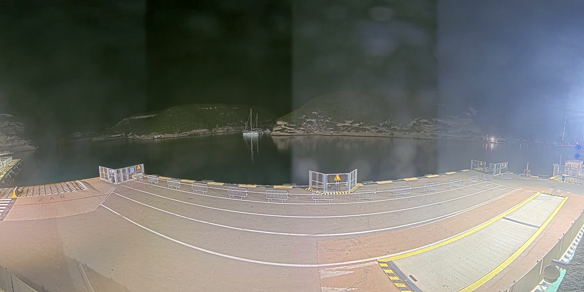 A nighttime view of a harbor at a ferry terminal with a sailboat in the water and barriers on the dock under a dark sky.