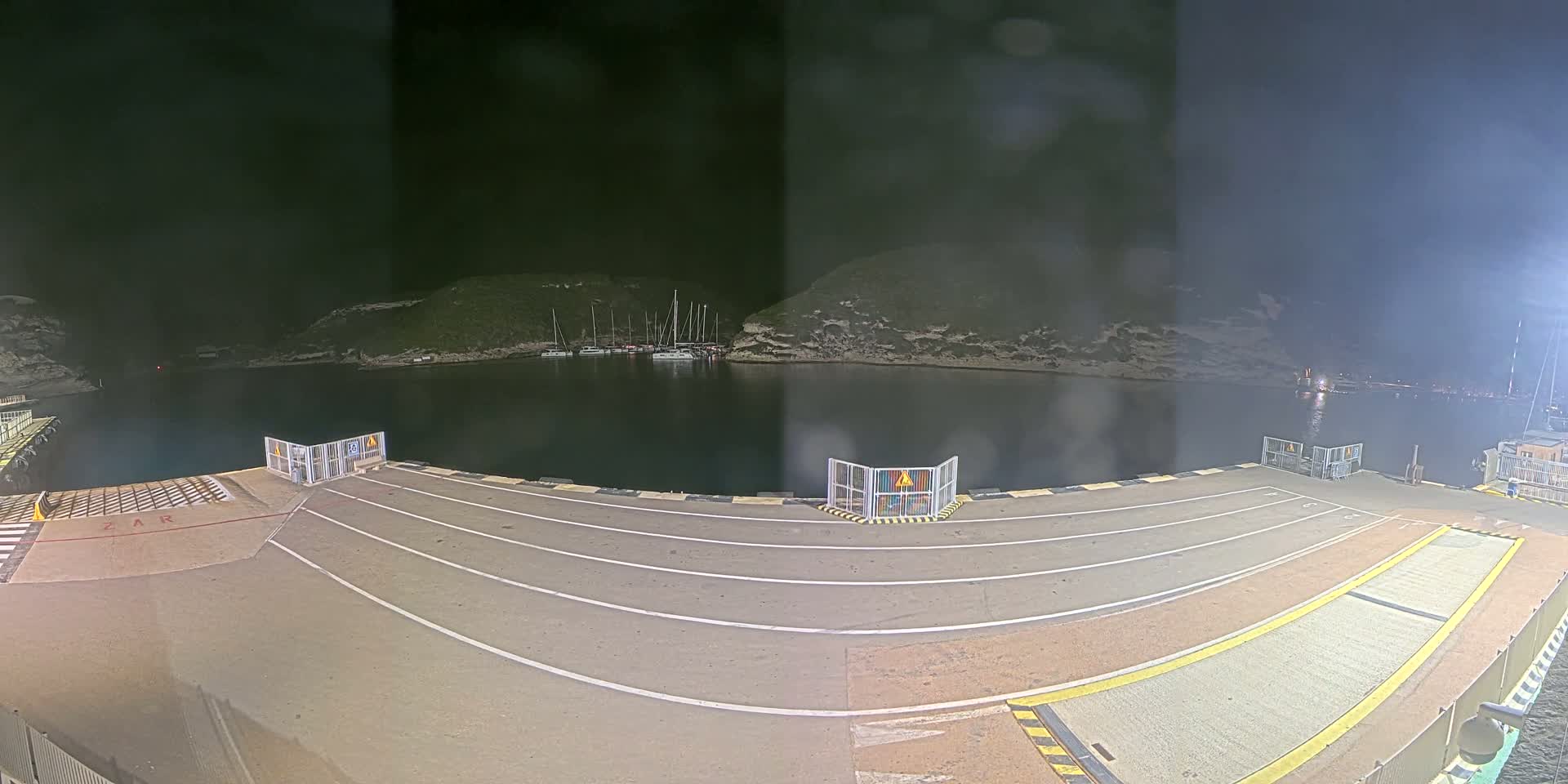 A nighttime view of a ferry terminal with several boats moored in a calm, dark bay, surrounded by hills.