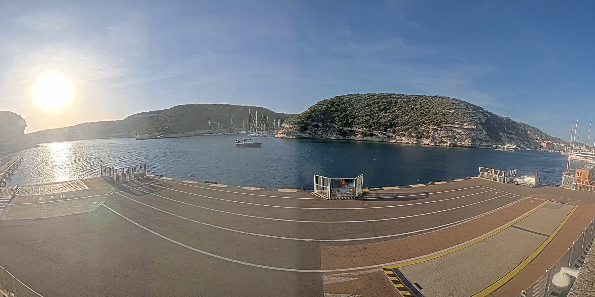 A sunny day at a harbor with calm water, boats moored in a cove surrounded by rocky hills.