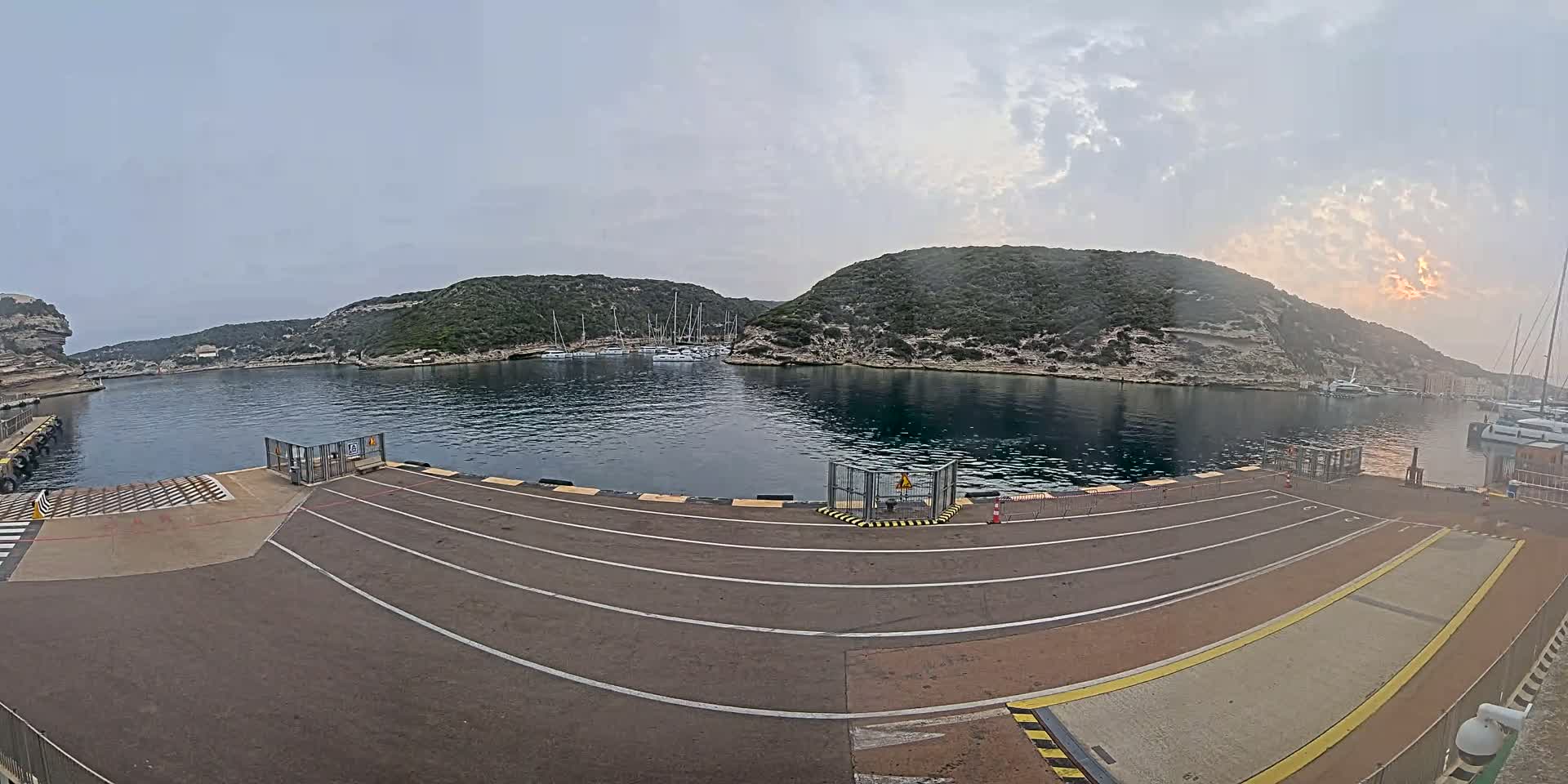 A harbor with calm water, surrounded by rocky hills covered in green vegetation, under a partly cloudy sky.