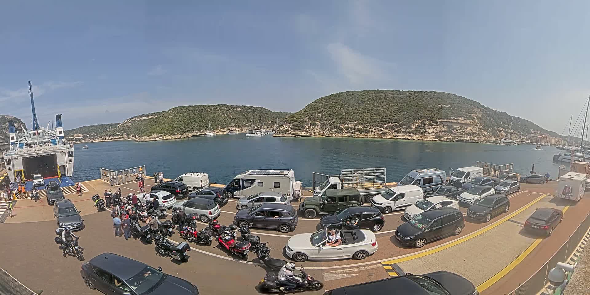 A ferry is docked at a port on a sunny day, surrounded by numerous cars, motorcycles, and people, with green hills and calm water in the background.