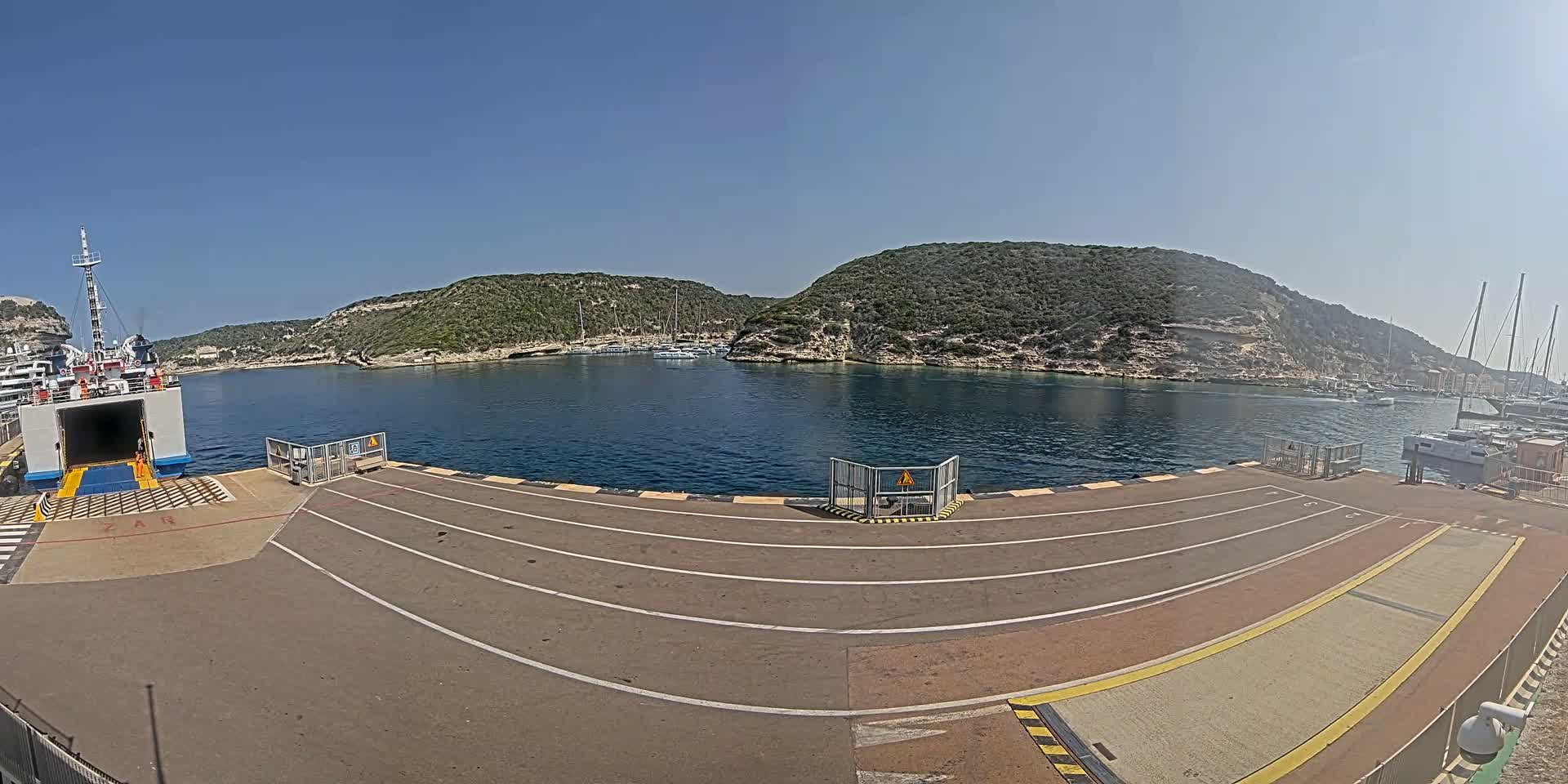 A ferry is docked at a port on a sunny day, with calm water and green hills in the background.